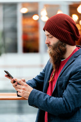 Handsome trendy hipster in hat and blue jacket using smartphone indoors