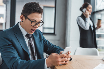 emotional young asian businessman using smartphone and female colleague talking behind in office