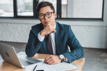 handsome young asian businessman in suit and eyeglasses sitting at workplace and smiling at camera