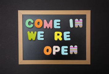Board with black frame, text Come in, we re open in colorful letters, black wall background