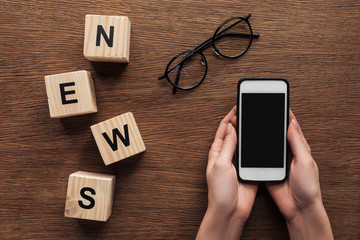 cropped image of journalist holding smartphone, wooden cubes with word news on wooden table