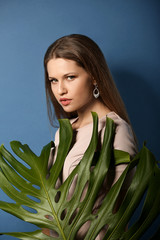 Naklejka premium Young woman with long hair and diamond earrings posing with large green monstera leaf on blue background, she looks directly at the camera with a serene expression