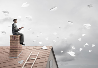 Man on brick roof reading book and paper planes flying in air