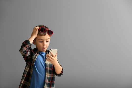 Emotional little boy playing with smartphone on grey background