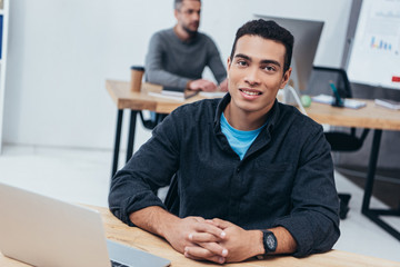 handsome young businessman sitting at workplace and smiling at camera