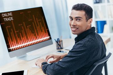 Fototapeta na wymiar young mixed race businessman using desktop computer with online trade charts and smiling at camera