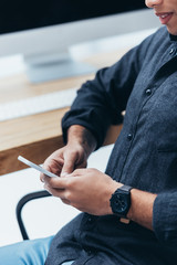 cropped shot of smiling young businessman sitting and using smartphone in office