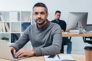 handsome man looking at camera while sitting at table with desktop computer in office