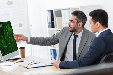 bearded businessman showing desktop computer with business graphs to young colleague at workplace