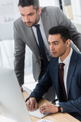 bearded business mentor looking at young colleague using desktop computer at workplace