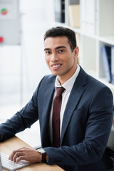 handsome young businessman working with desktop computer and smiling at camera in office
