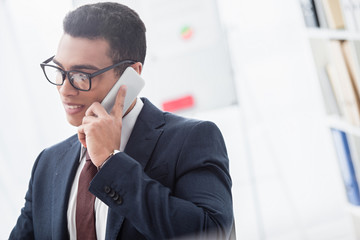 smiling young businessman in eyeglasses talking by smartphone and looking away in office