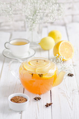 Hot fruit tea with fresh orange, lemon, anise, carnation and honey with ginger on a white wooden table. White porcelain cup, teapot with tea. White flowers. Different leaves. White porcelain bowl.