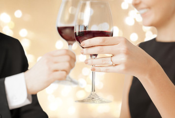 celebration, engagement and people concept - hands of couple with red wine in glasses and diamond ring over beige background with festive lights