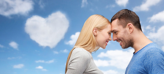 love, valentines day and people concept - smiling couple standing forehead to forehead over blue sky and heart shaped cloud background
