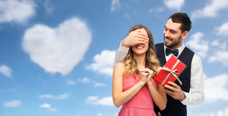 valentines day, couple, love and people concept - happy man giving woman surprise present over blue sky and heart shaped cloud background