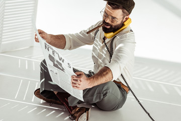 handsome bearded man sitting on floor, talking on phone, smoking cigar and reading business newspaper