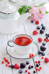 Berry red tea with fresh raspberries, currant, strawberries, blackberries and blueberries on a white wooden table. Fruit tea with fresh berries. White porcelain cup, teapot with tea. Pink white flower