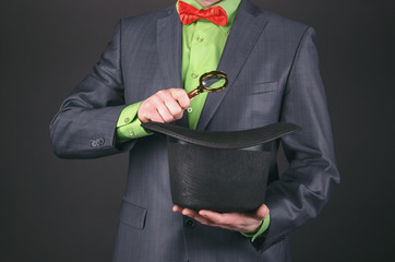 A magician is looking something in his hat by a magnifying glass on a black background.