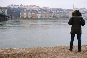 Man in winter jacket on river Danube bank in Budapest, Hungary. Buda bank from Pest side. Budapest landmark. Tourist on Danube bank in Budapest near bridge. Hungarian capital landscape. Travel concept