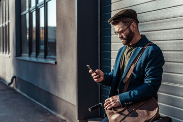 handsome bearded man in glasses and cap holding bicycle and looking at smartphone