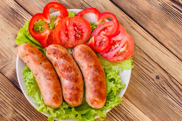 Ceramic plate with grilled sausages, sliced tomatoes and lettuce leaves on wooden table. Top view
