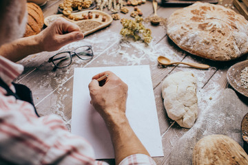 Old Baker writing down old-time recipe in bakery notebook surrounded by ingredients for making...