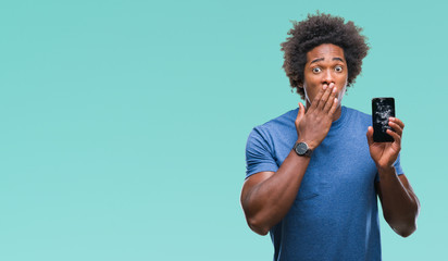 Afro american man holding broken smartphone over isolated background cover mouth with hand shocked...
