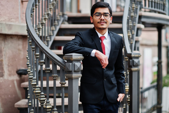 Indian young man at glasses, wear on black suit with red tie posed outdoor against iron stairs.