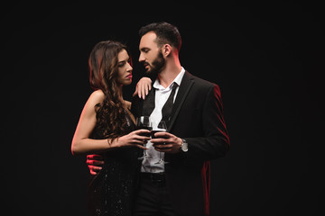 handsome boyfriend in tuxedo hugging girlfriend and holding glasses with red wine isolated on black