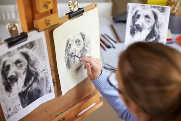 Rear View Of Female Teenage Artist Sitting At Easel Drawing Picture Of Dog From Photograph In...