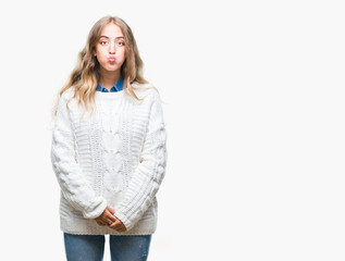 Beautiful young blonde woman wearing winter sweater over isolated background puffing cheeks with funny face. Mouth inflated with air, crazy expression.