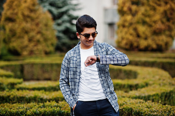 Stylish indian young man at sunglasses wear casual posed outdoor and looking at his watches on hand.