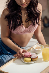 Obraz na płótnie Canvas cropped shot of sensual young woman in bra eating macarons for breakfast in bedroom