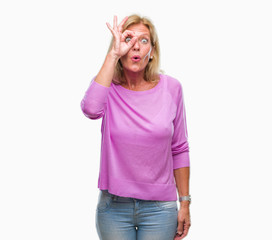 Middle age blonde woman over isolated background doing ok gesture shocked with surprised face, eye looking through fingers. Unbelieving expression.