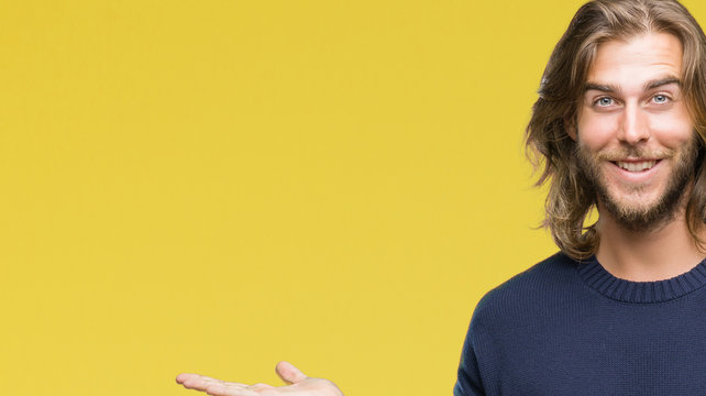 Young handsome man with long hair wearing winter sweater over isolated background amazed and smiling to the camera while presenting with hand and pointing with finger.