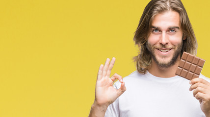 Young handsome man with long hair eating chocolate bar over isolated background doing ok sign with fingers, excellent symbol