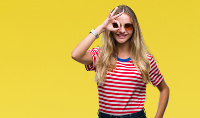 Young beautiful blonde woman wearing sunglasses over isolated background doing ok gesture with hand smiling, eye looking through fingers with happy face.
