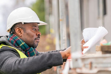 Profile portrait of an absorbed African American male engineer wearing safety jacket and helmet checking technical drawings and office blueprints among scaffolding