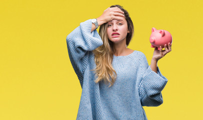 Young beautiful blonde woman holding piggy bank over isolated background stressed with hand on...