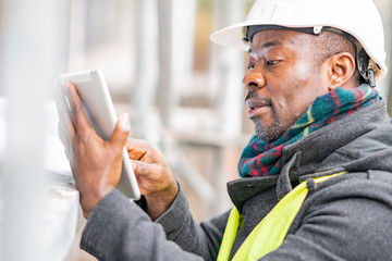 African American engineer wearing safety helmet and jacket checking documents on tablet computer on...