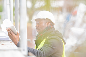 African American engineer wearing safety helmet and jacket checking documents on tablet computer on...