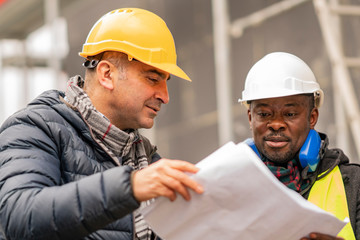 Civil engineers with hardhat and yellow jacket checking technical drawings and office blueprints...