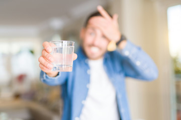 Young handsome man drinking a glass of water at home stressed with hand on head, shocked with shame and surprise face, angry and frustrated. Fear and upset for mistake.