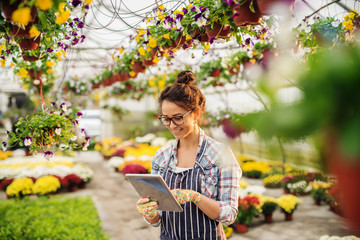 Young happy Caucasian female florist dressed in apron using tablet for selling flowers. Potted flowers all around.