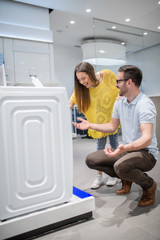 Couple laughing in front of washing machine. Man .squatting while woman leaning on him. Tech store interior.