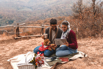 Couple at picnic sitting on the blanket and using tablet. Woman holding tablet while man holding tea and dog in his lap. In front of them basket. Autumn time.