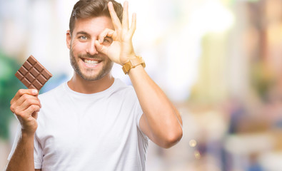 Young handsome man eating chocolate bar over isolated background with happy face smiling doing ok sign with hand on eye looking through fingers