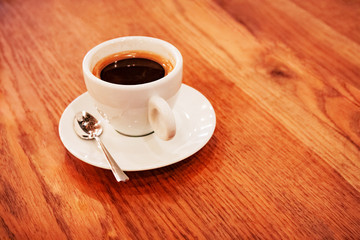 Close up white coffee cup with freshly brewed coffee and a spoon on a saucer stands on a dark wooden table.