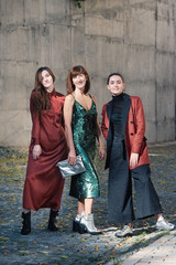 Street style Fashion portrait three charming women. Brown and green dress, Black, tile red jacket, silver shoes. Summer Spring Fall collection beautiful look. Togetherness, friendship, party concept